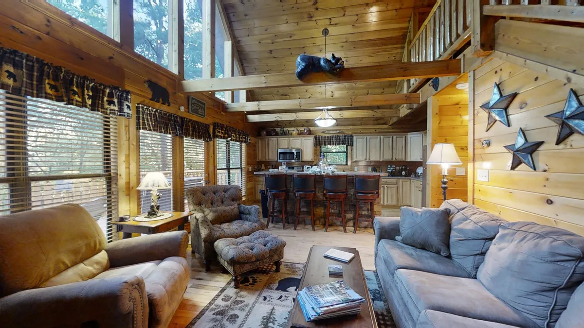 This image showcases a warm, welcoming living space with comfortable seating, expansive windows letting in natural light, visible wooden beams, and a kitchen island. The large windows subtly connect the indoors to the surrounding woods, hinting at the peaceful setting and inviting families to gather, relax, or plan their next adventure.