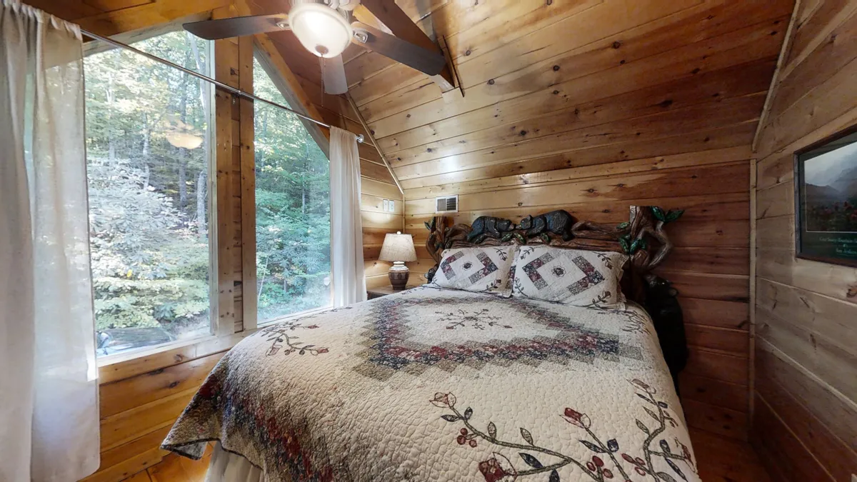 A warmly lit bedroom with a wooden ceiling, a large window letting in abundant natural light, and a quilted bedspread on the bed. The window offers a glimpse of nature, suggesting the tranquility and beauty of the Smokies just beyond the cabin walls.