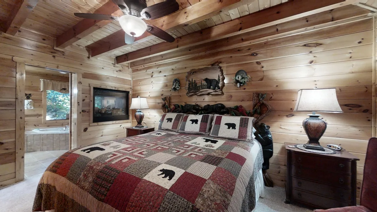 The image features a bed with a decorative quilt, wooden wall paneling, nightstands with lamps, and nature-themed artwork. The scene is cozy and exudes warmth, ideal for curling up and unwinding in the primary suite.