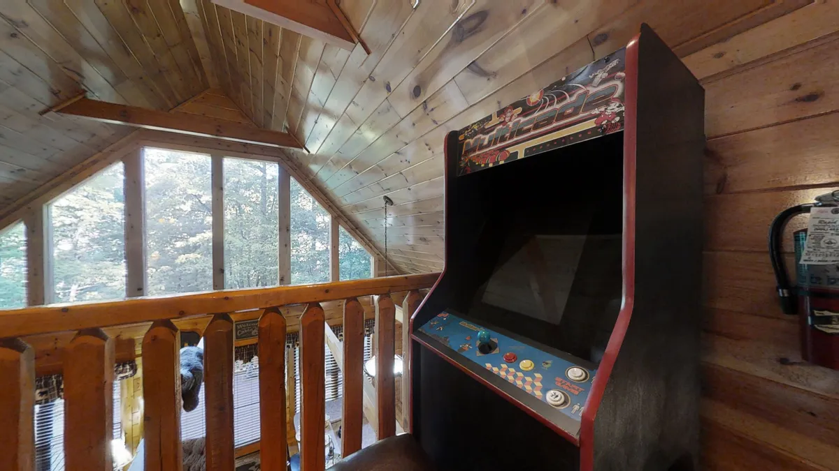 This image features an arcade machine set in a loft area with large windows and a wooden balcony visible, inviting both play and relaxation. The natural light streaming in and the elevated view add to the sense of fun and retreat. The playful mood is highlighted by the arcade machine, while the open layout hints at the multi-purpose nature of the space.