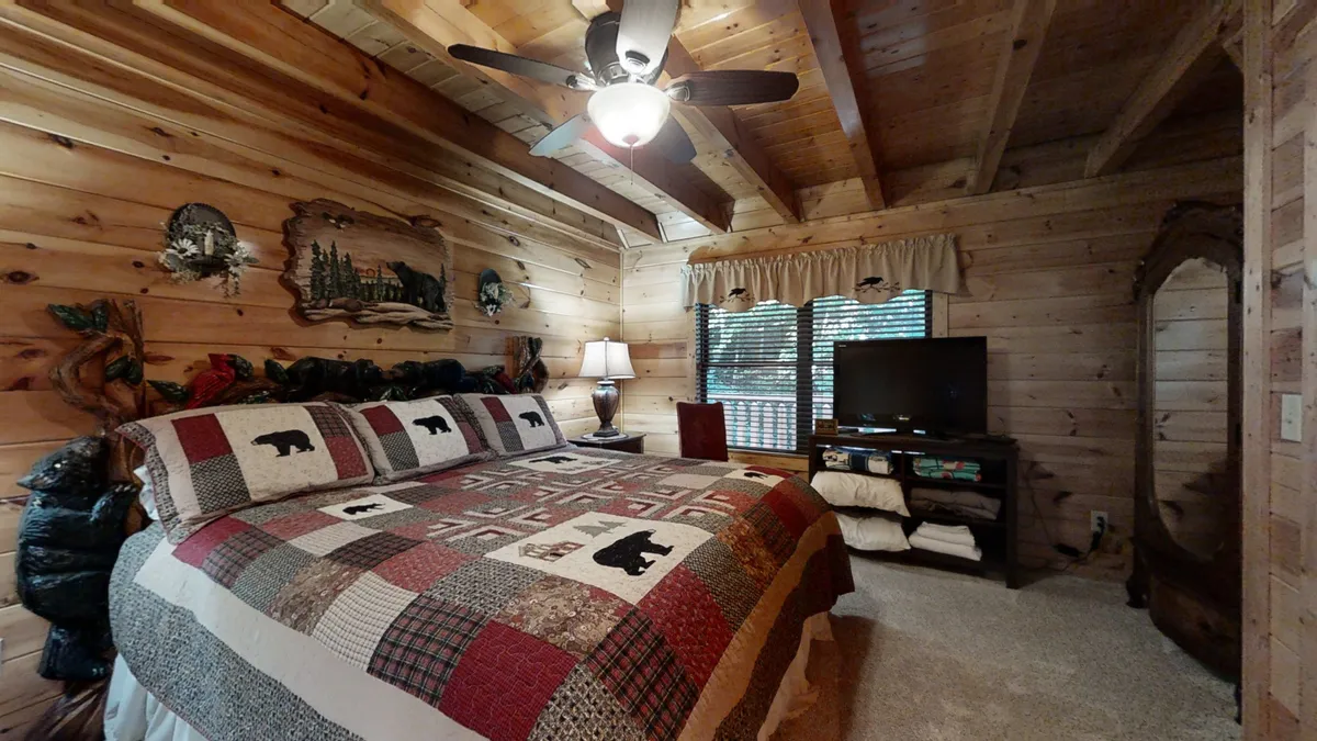 This image shows a bed with a bear-themed quilt in a rustic loft setting, wooden walls, a ceiling fan, a lamp, and a television. The cozy mood is accentuated by the soft lighting and inviting decor, making it ideal for relaxation, watching movies, or unwinding after activities.