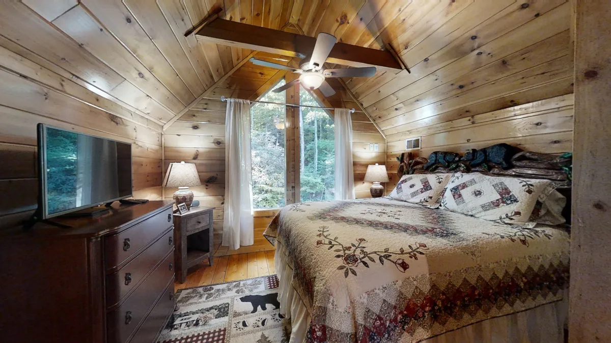 A cozy bedroom featuring a bed with a patterned quilt, wooden furniture, and a large window that opens up to a natural view. The window frames lush greenery, inviting the outdoors in. The room is tranquil and comfortable, blending interior warmth with visual access to the surrounding nature.