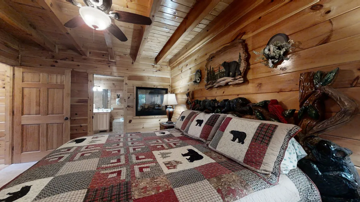 A warmly lit bedroom with bear-themed bedding, wooden wall art, a table lamp, and a fireplace. The setup invites couples to relax in a cozy ambiance, perfect for enjoying the warmth of the fire and the comfort of the room.