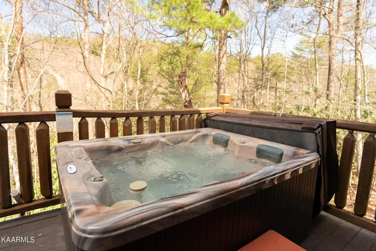 A serene view of the outdoor hot tub situated on the wooden wraparound porch, surrounded by tall trees and natural greenery. The deck's railing frames the setting, emphasizing the secluded, peaceful ambiance. The scene hints at early morning or dusk, inviting relaxation and connection with nature.