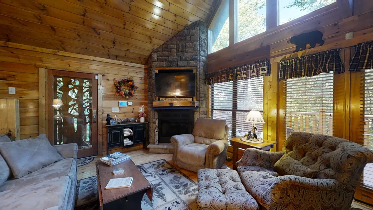 A bright and airy living room featuring a grand stone fireplace as the focal point, flanked by large windows that allow sunlight to pour into the space. Comfortable seating is arranged to encourage conversation and relaxation, and the room is accented by exposed wooden beams for a rustic yet inviting atmosphere.