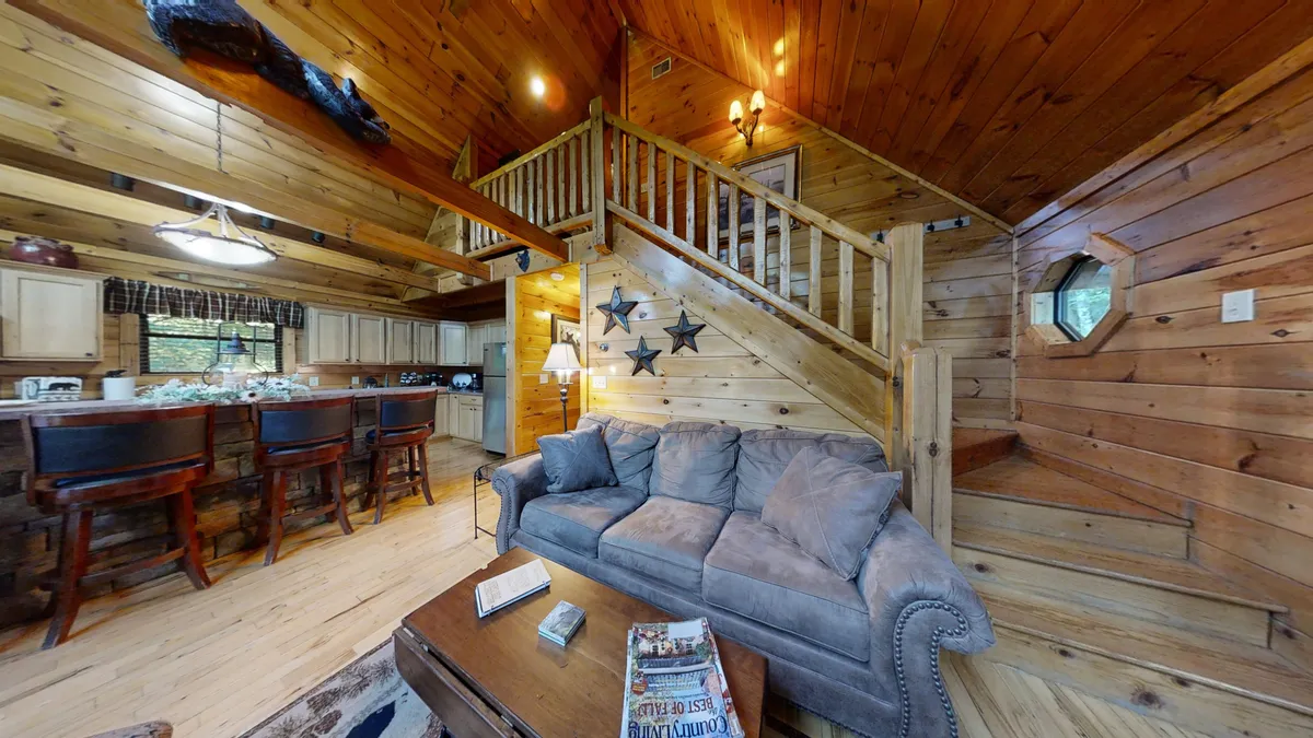 A seamless view into the open living area, showing a plush sofa, the kitchen bar area with stools, and a wooden staircase. Decorative elements like wall stars add charm while the layout highlights the connection between the kitchen and living room, emphasizing the open flow ideal for gatherings.
