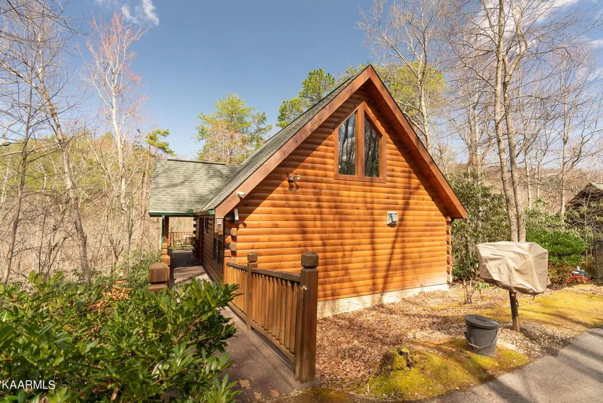 This exterior image features a charming wooden cabin surrounded by lush green foliage, set amidst the tranquil Smoky Mountain woods. The porch wraps around the cabin, inviting guests to enjoy the peaceful outdoor environment and immerse themselves in nature.