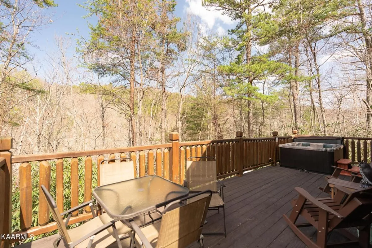 A wider exterior perspective of the wraparound deck, featuring not only the hot tub but also outdoor furniture like a table and chairs. The scene is framed by the surrounding trees and hints at mountain views beyond, inviting gatherings and morning coffee outdoors.