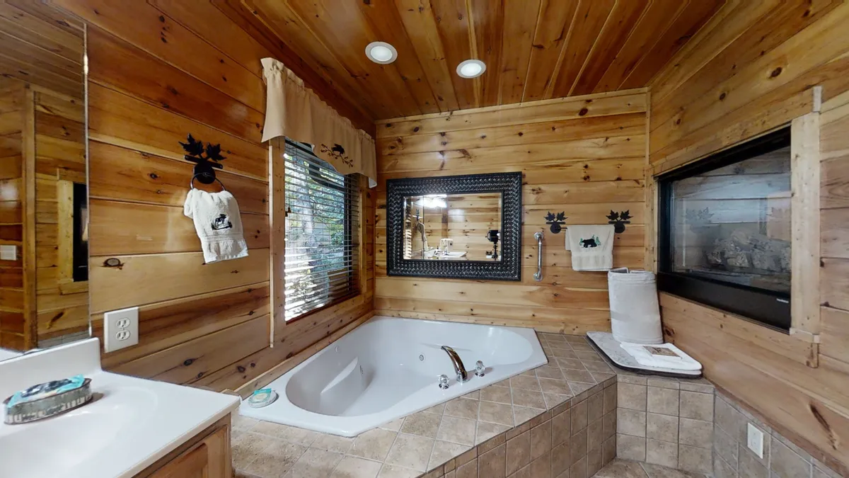 This image showcases a jacuzzi tub set against warm wooden paneling, with a large mirror above and a window providing natural light and views of the outdoors. The inviting setting is ideal for a relaxing soak and features tranquil decorative elements.